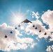 A dramatic aerial formation photograph taken from below, showing a large flying-wing stealth bomber (consistent with a B-2 Spirit) flanked by multiple smaller twin-tail fighter jets (consistent with F-22 Raptors) flying in a V-shaped escort formation. The image is backlit by a bright sunburst against a blue sky with large cumulus clouds, rendering all aircraft as dark silhouettes. The composition creates a striking contrast between the aircraft silhouettes and the luminous sky.