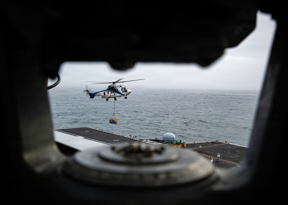 A white and blue helicopter, appearing to be a medium-lift utility type (likely an Aerospatiale/Eurocopter AS332 Super Puma), is conducting a vertical replenishment (VERTREP) operation over the flight deck of a naval vessel. The helicopter is carrying a sling-loaded cargo package suspended beneath it, while deck personnel in various colored jerseys stand by on the flight deck. The image is captured through a circular hatch or porthole opening on the ship, framing the scene dramatically against an overcast sea background.
