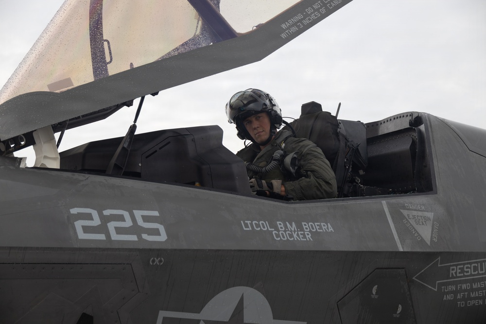 A military pilot is seated in the cockpit of a stealth fighter jet, identified by markings as aircraft number 225, assigned to Lt Col B.M. Boera with the callsign 'Cocker'. The aircraft appears to be an F-35 based on its distinctive stealth design and cockpit configuration. The canopy is open and the pilot is wearing full flight gear including a helmet and oxygen mask connection.