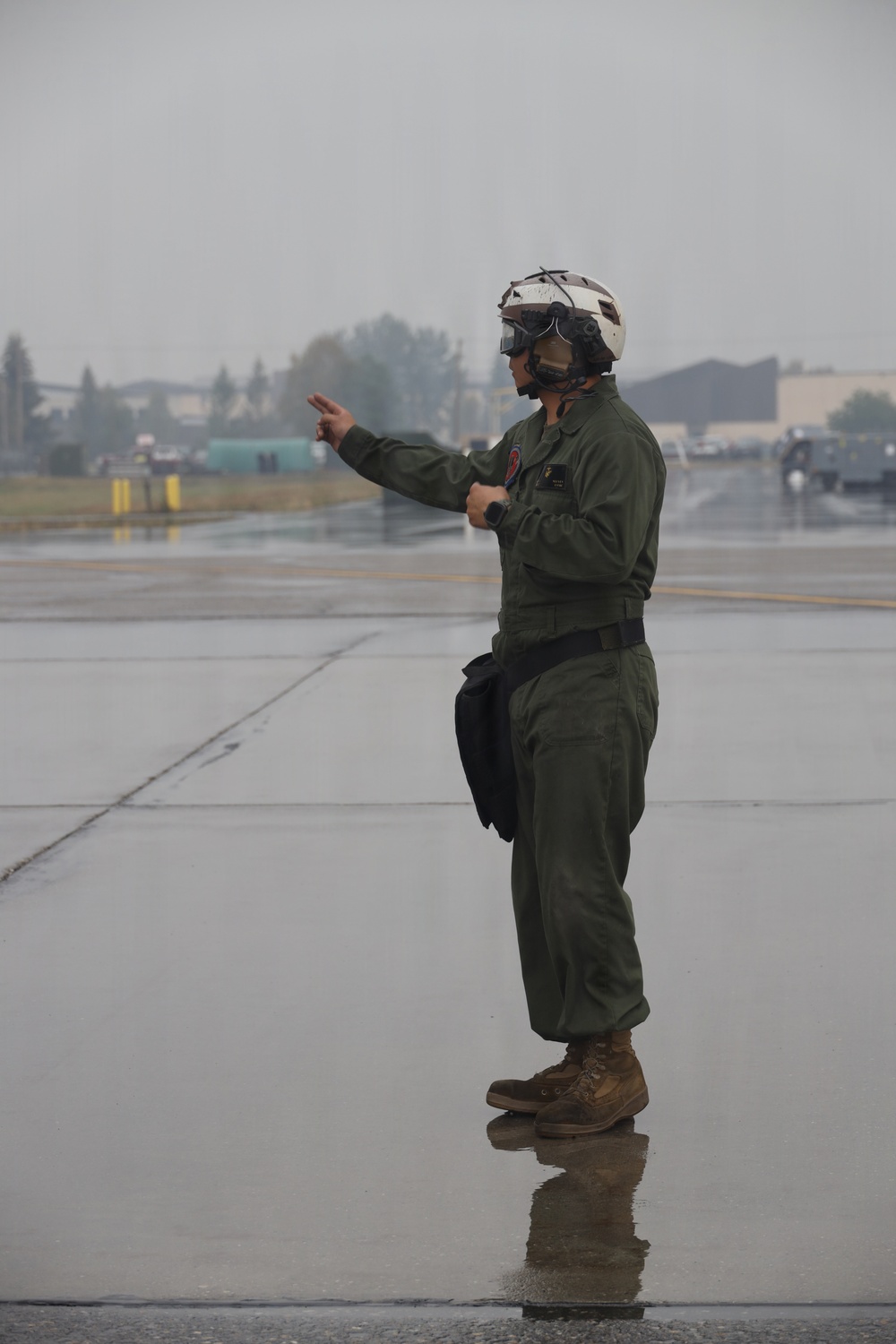 A military flight crew member stands on a wet tarmac/flight line, wearing olive green flight suit and a white flight helmet with visor. The individual is gesturing with one arm extended outward, likely performing aircraft marshaling or directing duties. The weather conditions appear overcast and rainy, with a reflective wet surface visible beneath their boots.