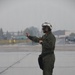 A military flight crew member stands on a wet tarmac/flight line, wearing olive green flight suit and a white flight helmet with visor. The individual is gesturing with one arm extended outward, likely performing aircraft marshaling or directing duties. The weather conditions appear overcast and rainy, with a reflective wet surface visible beneath their boots.