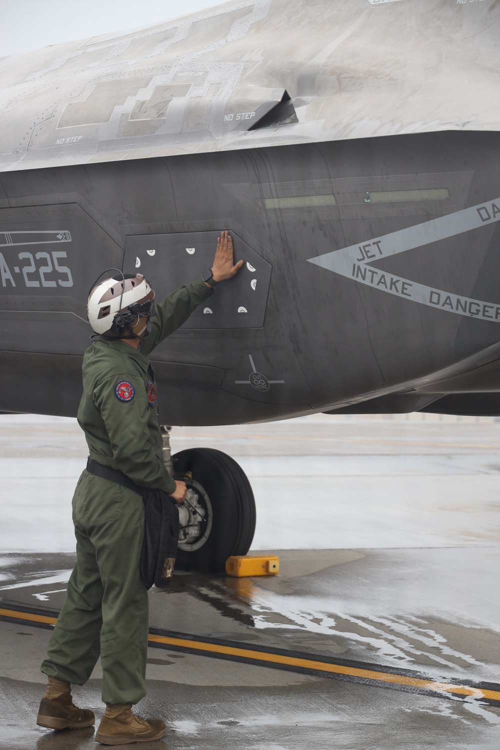 A military aviation crew member in a green flight suit and white helmet is conducting a pre-flight or post-flight inspection of a stealth fighter aircraft, identified by the tail code 'A-225'. The aircraft displays markings including 'JET INTAKE DANGER' warning text and 'NO STEP' labels, consistent with an F-35 Lightning II. The scene takes place on a wet tarmac, suggesting recent rainfall.