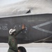 A military aviation crew member in a green flight suit and white helmet is conducting a pre-flight or post-flight inspection of a stealth fighter aircraft, identified by the tail code 'A-225'. The aircraft displays markings including 'JET INTAKE DANGER' warning text and 'NO STEP' labels, consistent with an F-35 Lightning II. The scene takes place on a wet tarmac, suggesting recent rainfall.