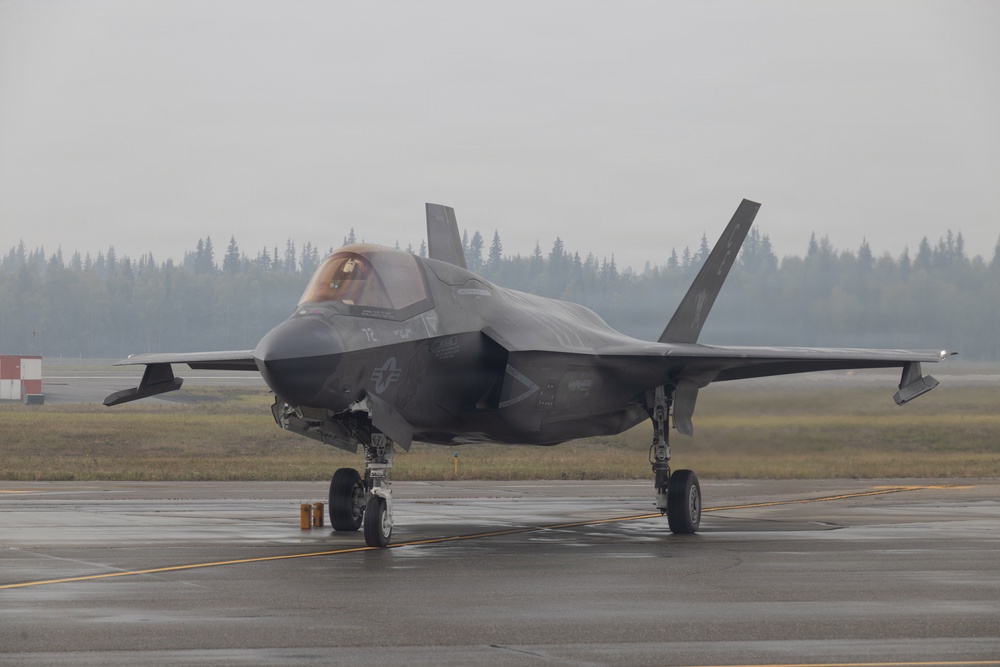 A single-seat stealth fighter jet, identifiable as an F-35 Lightning II, is parked on a wet tarmac under overcast, hazy conditions. The aircraft bears U.S. military markings including a star-and-bar insignia on the fuselage and the number '72' near the cockpit. A pilot is visible in the cockpit, and the aircraft appears to be on the ground with landing gear deployed.