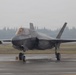 A single-seat stealth fighter jet, identifiable as an F-35 Lightning II, is parked on a wet tarmac under overcast, hazy conditions. The aircraft bears U.S. military markings including a star-and-bar insignia on the fuselage and the number '72' near the cockpit. A pilot is visible in the cockpit, and the aircraft appears to be on the ground with landing gear deployed.