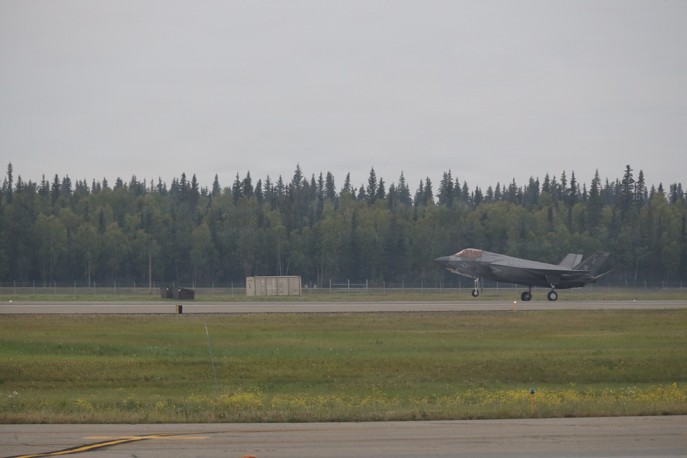 A single F-35 Lightning II stealth fighter jet is taxiing or landing on a runway at a military airfield. The aircraft appears to be in a low-speed ground roll with its canopy open and landing gear deployed. The setting appears to be a northern or Alaskan airbase, suggested by the dense coniferous treeline in the background.