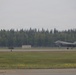 A single F-35 Lightning II stealth fighter jet is taxiing or landing on a runway at a military airfield. The aircraft appears to be in a low-speed ground roll with its canopy open and landing gear deployed. The setting appears to be a northern or Alaskan airbase, suggested by the dense coniferous treeline in the background.