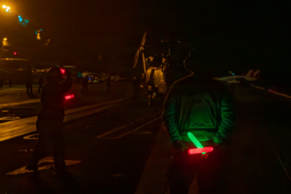 A nighttime scene on what appears to be a naval aircraft carrier flight deck, with deck crew members using illuminated wands to guide aircraft operations. Two personnel are prominently visible in the foreground holding colored light wands (green and red), consistent with standard flight deck signaling procedures. A helicopter with spinning rotors and what appears to be a fixed-wing aircraft are visible in the background.