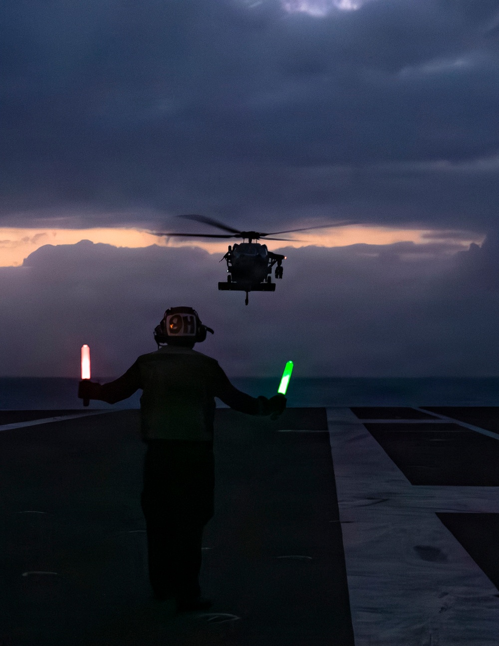 A military flight deck handler stands on a ship's flight deck at dusk, guiding a helicopter toward landing using illuminated signal wands. The handler holds a red wand in the left hand and a green wand in the right hand, facing a hovering military helicopter silhouetted against a dramatic twilight sky. The scene depicts a nighttime or low-light naval flight deck operations procedure.