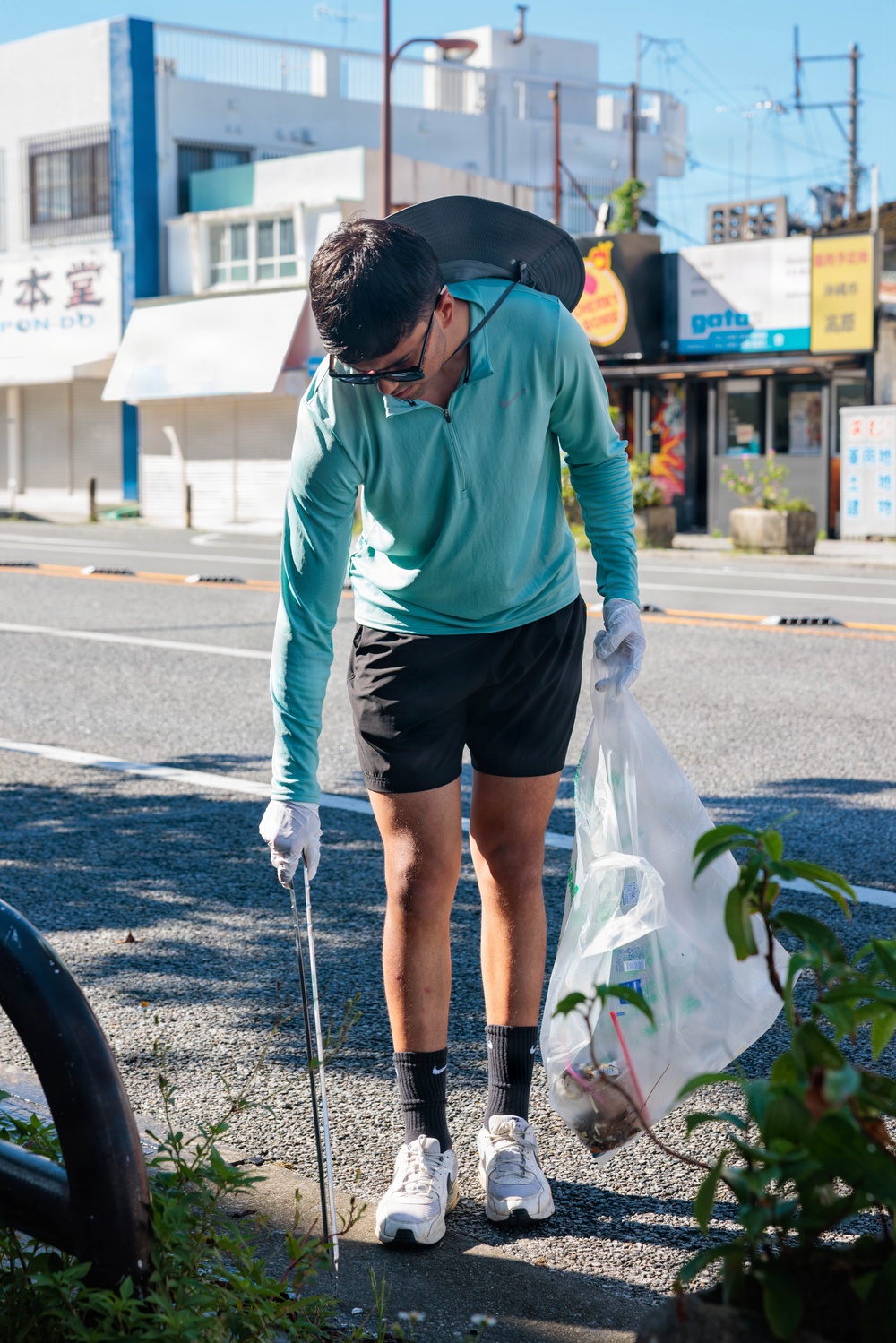 A person is engaged in a street litter-picking activity along a Japanese urban road. They are bent forward using a litter picker tool while holding a clear plastic collection bag, wearing protective gloves, sunglasses, and a wide-brimmed hat pushed back on their head. The background shows Japanese commercial storefronts and street infrastructure under clear blue skies.