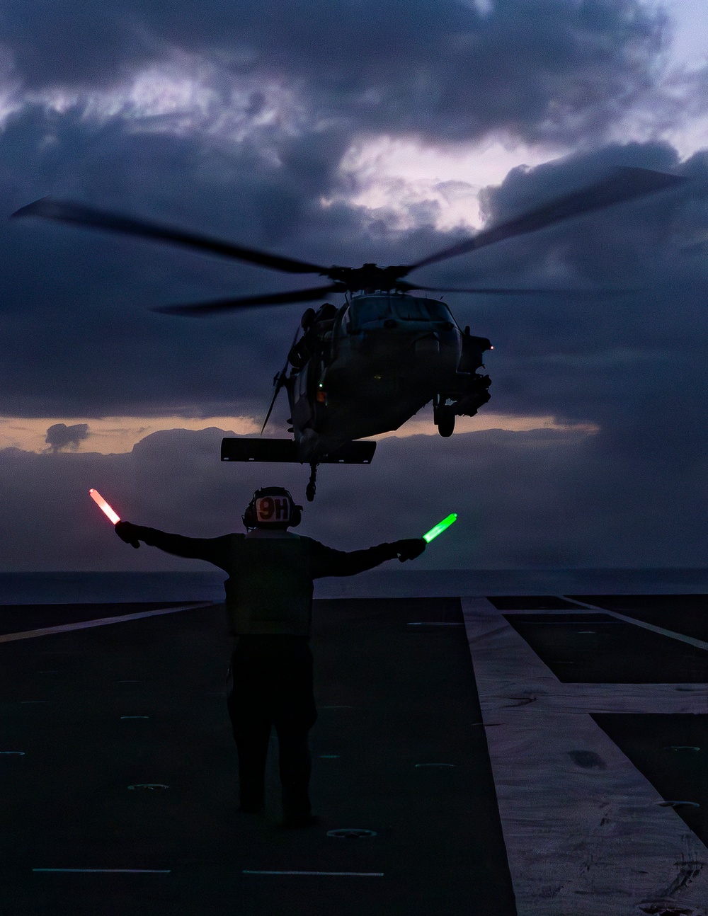A naval flight deck crew member guides a military helicopter during a dusk or dawn landing operation aboard a ship at sea. The handler holds illuminated red and green marshalling wands with arms outstretched, directing the hovering helicopter. The scene is captured in low-light conditions with dramatic cloud cover over open ocean.