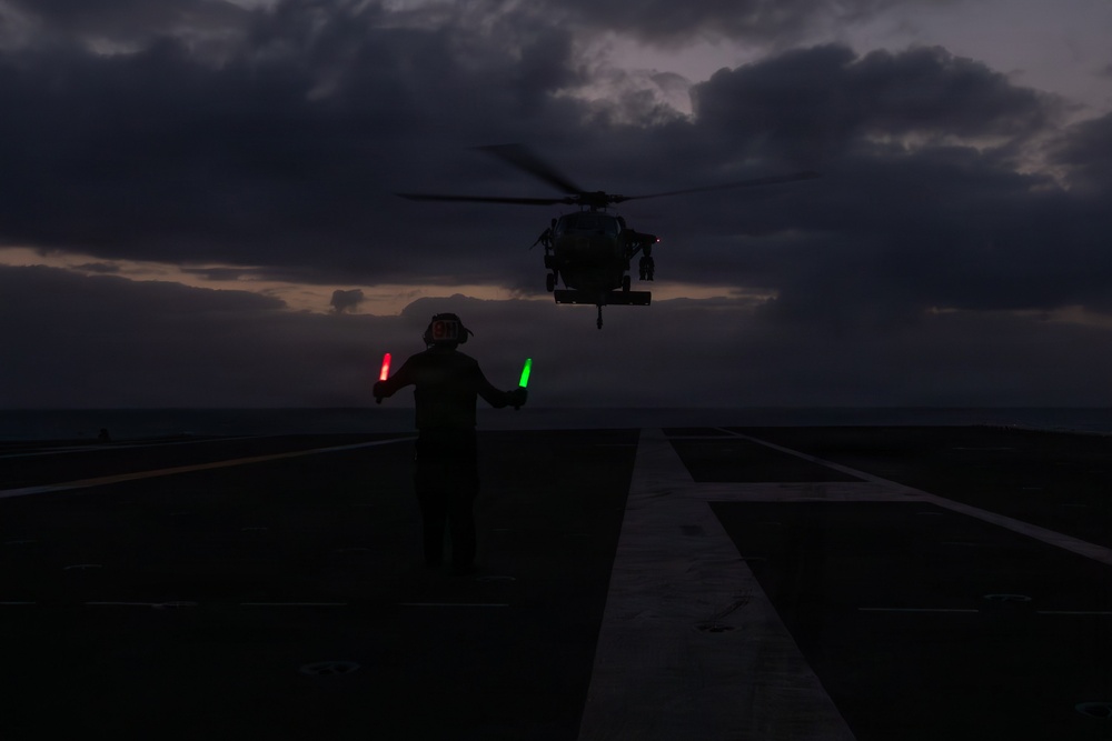 A flight deck crew member guides a military helicopter during a dusk or nighttime landing operation on what appears to be a naval vessel. The handler is using illuminated signal wands — one red and one green — to direct the aircraft. The scene is dramatically backlit by a dark, cloudy sky with faint twilight on the horizon.