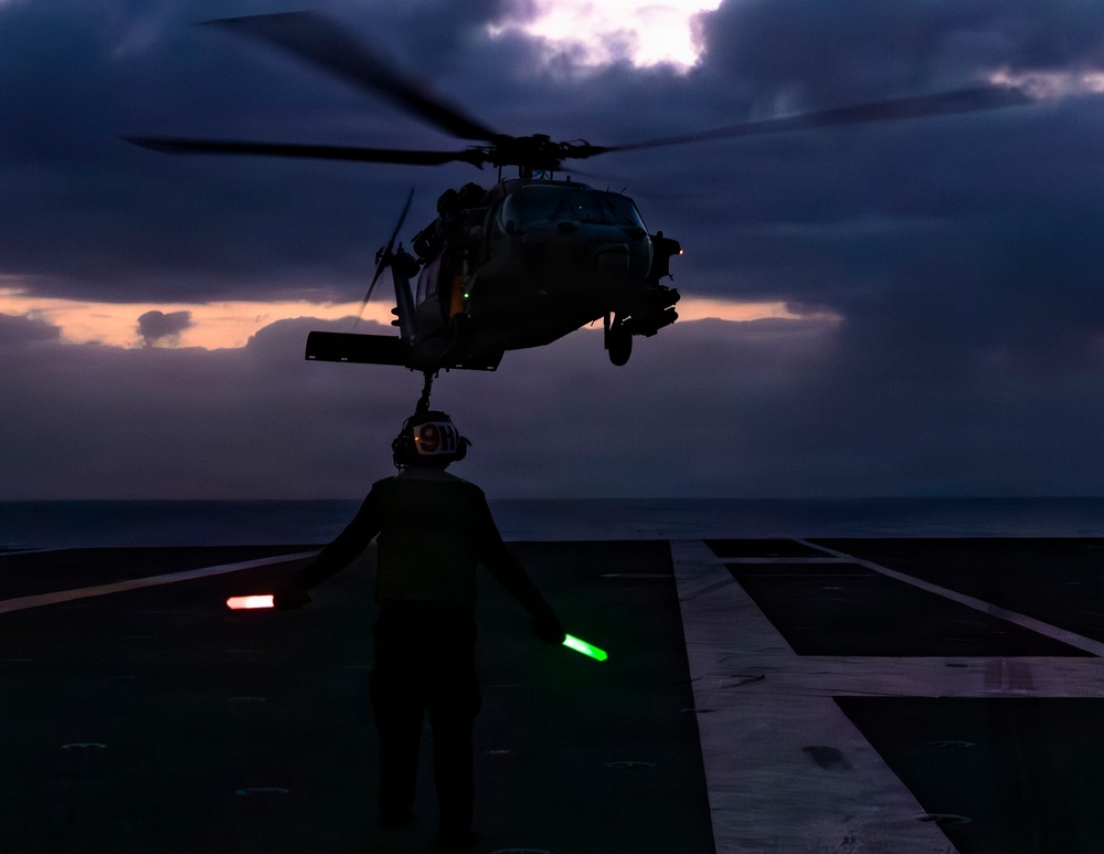 A military helicopter, appearing to be an MH-60 Seahawk variant, hovers over a naval vessel's flight deck at dusk or dawn. A deck crew member stands beneath the aircraft holding a green light wand, guiding the helicopter during landing or hover operations. The dramatic low-light conditions create a silhouetted scene against a stormy, cloud-filled sky over open ocean.