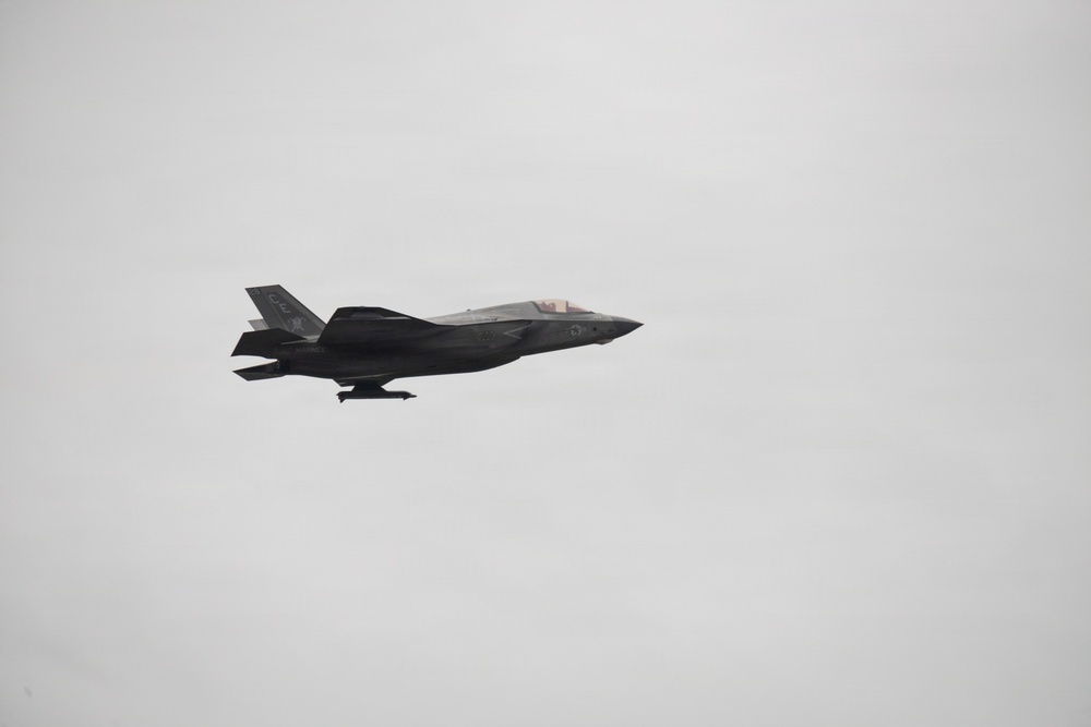 A single stealth fighter jet is captured in flight against a uniformly overcast gray sky. The aircraft displays characteristics consistent with an F-35 Lightning II, featuring a dark gray paint scheme, single engine configuration, and distinctive stealth airframe design. The aircraft appears to be in level flight with landing gear retracted.