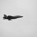 A single stealth fighter jet is captured in flight against a uniformly overcast gray sky. The aircraft displays characteristics consistent with an F-35 Lightning II, featuring a dark gray paint scheme, single engine configuration, and distinctive stealth airframe design. The aircraft appears to be in level flight with landing gear retracted.