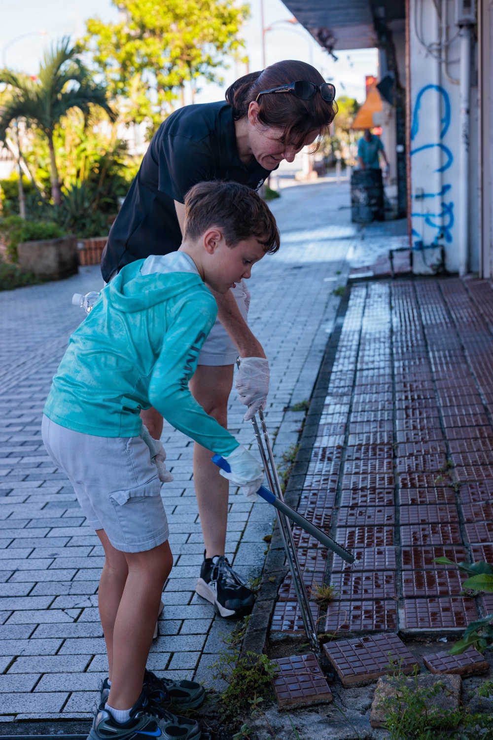 A woman and a young boy are engaged in a community cleanup activity on a sidewalk, using grabber/reacher tools and wearing protective gloves. They are bent over picking up debris or weeds from the edge of a tiled walkway along a commercial street. A third person is visible in the background performing a similar activity.