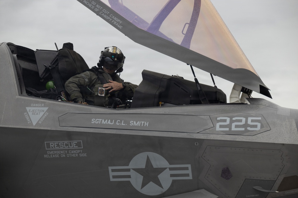 A military pilot sits in the open cockpit of an F-35 fighter jet, wearing full flight gear including helmet and oxygen mask. The aircraft bears the name 'SGTMAJ C.L. SMITH' and the number '225' on its fuselage, along with standard US military markings including a star-and-bar insignia. The canopy is raised and the pilot appears to be conducting pre-flight or post-flight checks.