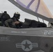 A military pilot sits in the open cockpit of an F-35 fighter jet, wearing full flight gear including helmet and oxygen mask. The aircraft bears the name 'SGTMAJ C.L. SMITH' and the number '225' on its fuselage, along with standard US military markings including a star-and-bar insignia. The canopy is raised and the pilot appears to be conducting pre-flight or post-flight checks.
