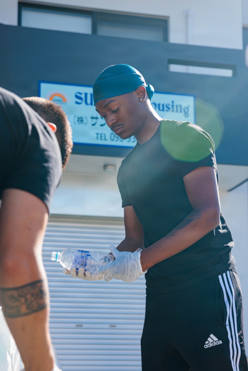 A young man wearing a teal durag, dark green t-shirt, and black Adidas track pants is holding a crushed plastic water bottle while wearing disposable gloves. He appears to be engaged in a cleanup or waste collection activity outdoors. A second person is partially visible on the left side of the frame, and a Japanese business sign reading 'Sun Housing' is visible in the background.