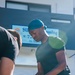 A young man wearing a teal durag, dark green t-shirt, and black Adidas track pants is holding a crushed plastic water bottle while wearing disposable gloves. He appears to be engaged in a cleanup or waste collection activity outdoors. A second person is partially visible on the left side of the frame, and a Japanese business sign reading 'Sun Housing' is visible in the background.