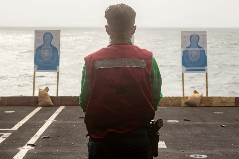 A person wearing a red vest over a green long-sleeve shirt stands on what appears to be a ship's deck facing two blue humanoid silhouette shooting targets. Spent brass casings are visible scattered on the deck surface, indicating a firearms qualification or training exercise has taken place. The ocean is visible in the background, confirming the maritime setting.