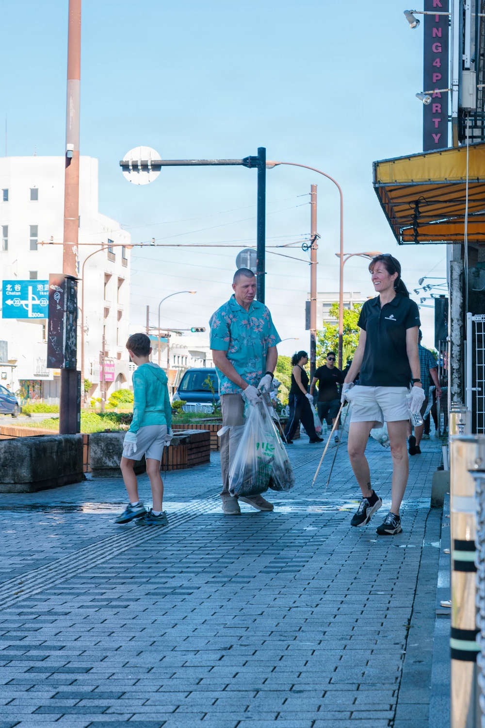 A group of people are participating in a community street cleanup along a sidewalk in what appears to be Okinawa, Japan, based on the visible Japanese road signs. Two adults in the foreground wear gloves and carry trash bags and a litter-picking stick, while a child and other participants are visible in the background. The setting is an urban commercial street with Japanese signage and infrastructure.