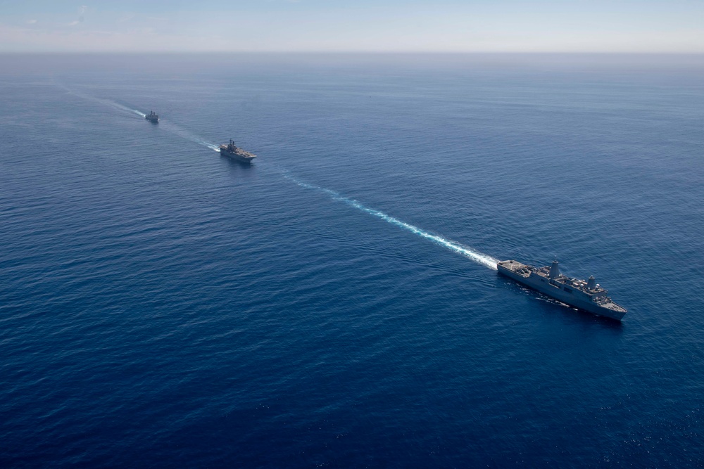 An aerial photograph shows three naval vessels sailing in a single-file formation across open, deep-blue ocean water. The ships are traveling in the same direction, leaving a visible white wake trail connecting them in a diagonal line across the frame. The formation appears to be a military naval exercise or transit operation.