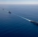 An aerial photograph shows three naval vessels sailing in a single-file formation across open, deep-blue ocean water. The ships are traveling in the same direction, leaving a visible white wake trail connecting them in a diagonal line across the frame. The formation appears to be a military naval exercise or transit operation.