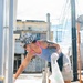 A tattooed individual wearing a camouflage bucket hat, blue tank top, and white gloves is bent over picking up litter on an urban street. They are holding a clear plastic garbage bag and appear to be participating in a street or neighborhood cleanup activity. The setting appears to be a Japanese urban environment based on visible infrastructure and signage.