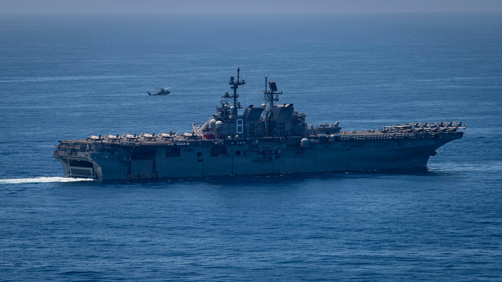 A large U.S. Navy amphibious assault ship bearing the hull number '8' is underway on open blue ocean waters. The vessel has a flat flight deck loaded with multiple aircraft, and a helicopter is visible in flight nearby. The ship appears to be the USS Makin Island (LHD-8) based on the hull number.