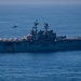 A large U.S. Navy amphibious assault ship bearing the hull number '8' is underway on open blue ocean waters. The vessel has a flat flight deck loaded with multiple aircraft, and a helicopter is visible in flight nearby. The ship appears to be the USS Makin Island (LHD-8) based on the hull number.