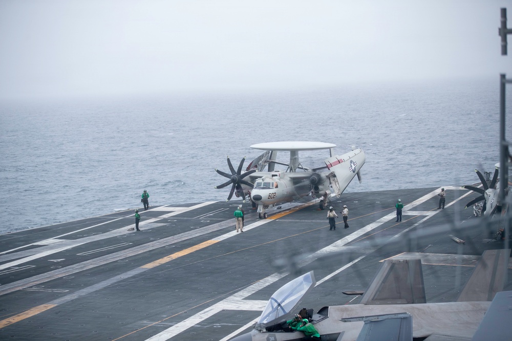 A U.S. Navy E-2 Hawkeye airborne early warning aircraft (bureau number 602) is positioned on the flight deck of an aircraft carrier, with its wings folded. Multiple flight deck crew members in green jerseys and white helmets are visible performing pre-flight or positioning operations. The scene is overcast with low visibility conditions over open ocean.
