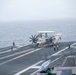 A U.S. Navy E-2 Hawkeye airborne early warning aircraft (bureau number 602) is positioned on the flight deck of an aircraft carrier, with its wings folded. Multiple flight deck crew members in green jerseys and white helmets are visible performing pre-flight or positioning operations. The scene is overcast with low visibility conditions over open ocean.