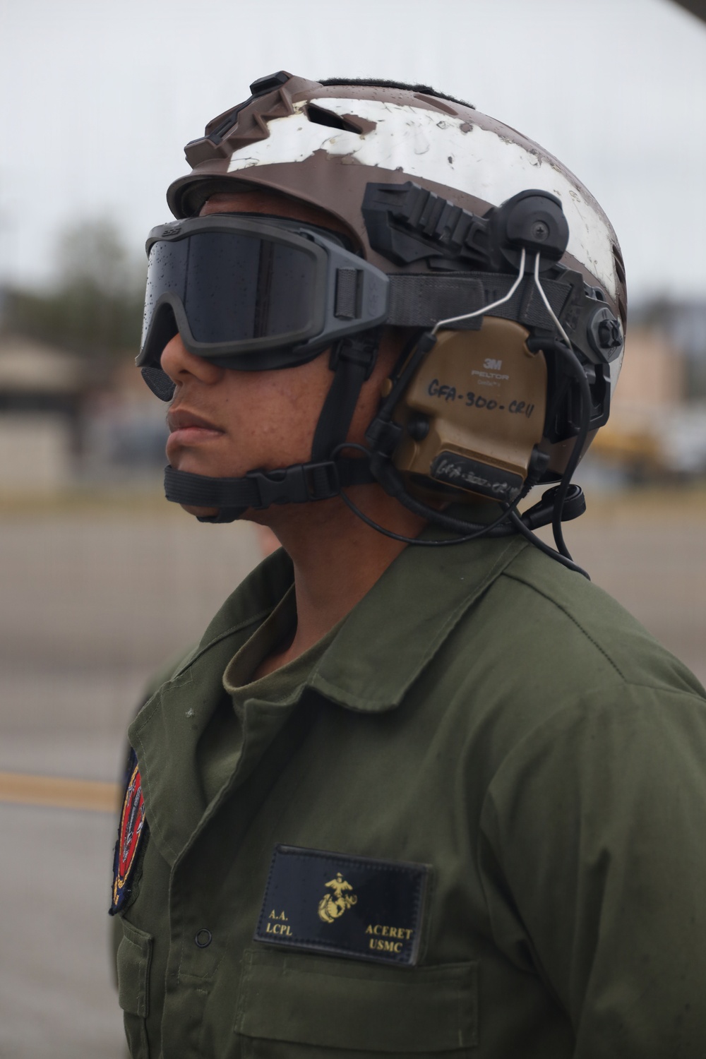 A United States Marine Corps Lance Corporal named Aceret is photographed in profile view on what appears to be a flight line or tarmac. The individual is wearing full flight deck/airfield safety gear including a tactical helmet with communications equipment and protective goggles. The name tape and rank insignia are clearly visible on the olive drab flight suit.