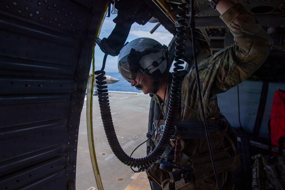 A military aircrew member is positioned at the open door of a helicopter, wearing a flight helmet with visor, multicam/OCP uniform, and connected to the aircraft via a coiled communication cord and safety tether. In the background, what appears to be a naval vessel deck is visible along with open ocean and sky. The image captures an in-flight or on-deck operational moment from inside the aircraft.
