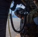 A military aircrew member is positioned at the open door of a helicopter, wearing a flight helmet with visor, multicam/OCP uniform, and connected to the aircraft via a coiled communication cord and safety tether. In the background, what appears to be a naval vessel deck is visible along with open ocean and sky. The image captures an in-flight or on-deck operational moment from inside the aircraft.