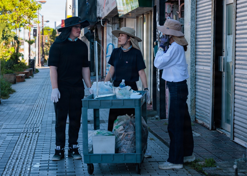 Three individuals are standing on a sidewalk next to a utility cart filled with collected trash and recyclables. They appear to be engaged in a community cleanup or waste collection activity, wearing sun hats and gloves for protection. The setting appears to be a commercial street in Japan, with shuttered storefronts visible in the background.