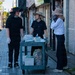 Three individuals are standing on a sidewalk next to a utility cart filled with collected trash and recyclables. They appear to be engaged in a community cleanup or waste collection activity, wearing sun hats and gloves for protection. The setting appears to be a commercial street in Japan, with shuttered storefronts visible in the background.