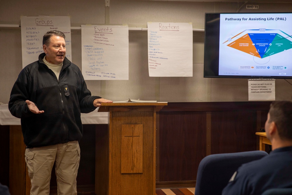 A man wearing a black fleece jacket with a cross pin is presenting at a podium in what appears to be a military chapel or training room. The presentation focuses on the 'Pathway for Assisting Life (PAL)' program, which appears to be a suicide prevention or mental health support training. Flip chart papers on the wall display brainstormed lists under headings 'Groups,' 'Events,' and 'Reactions,' suggesting an interactive training session.