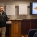 A man wearing a black fleece jacket with a cross pin is presenting at a podium in what appears to be a military chapel or training room. The presentation focuses on the 'Pathway for Assisting Life (PAL)' program, which appears to be a suicide prevention or mental health support training. Flip chart papers on the wall display brainstormed lists under headings 'Groups,' 'Events,' and 'Reactions,' suggesting an interactive training session.