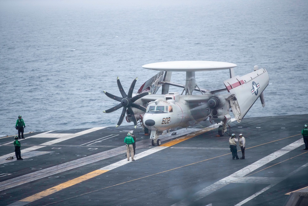 A U.S. Navy E-2 Hawkeye airborne early warning aircraft (bureau number 602) is positioned on the flight deck of an aircraft carrier at sea. The aircraft features its distinctive rotodome radar dish on top and has its wings folded for deck storage. Several flight deck crew members in green jerseys and white jerseys are visible attending to the aircraft in overcast, hazy conditions.