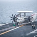A U.S. Navy E-2 Hawkeye airborne early warning aircraft (bureau number 602) is positioned on the flight deck of an aircraft carrier at sea. The aircraft features its distinctive rotodome radar dish on top and has its wings folded for deck storage. Several flight deck crew members in green jerseys and white jerseys are visible attending to the aircraft in overcast, hazy conditions.