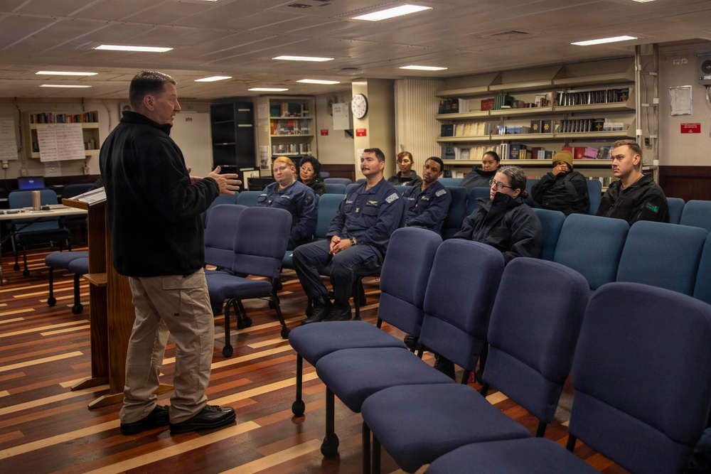 A civilian or senior official in casual attire addresses a small group of uniformed military personnel in what appears to be a briefing room or library aboard a naval vessel or military facility. The audience consists of approximately 8-9 individuals wearing dark blue military uniforms, seated in blue upholstered chairs. The setting includes bookshelves lined with reference materials along the back wall.