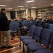 A civilian or senior official in casual attire addresses a small group of uniformed military personnel in what appears to be a briefing room or library aboard a naval vessel or military facility. The audience consists of approximately 8-9 individuals wearing dark blue military uniforms, seated in blue upholstered chairs. The setting includes bookshelves lined with reference materials along the back wall.