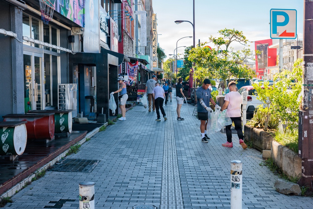 A group of people are conducting a community street cleanup along a urban sidewalk in what appears to be Japan, based on visible Japanese signage and architectural style. Multiple volunteers are walking along the paved pedestrian path carrying plastic bags, collecting litter. The scene takes place during daytime on a commercial street lined with shops, graffiti murals, and urban vegetation.