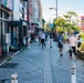 A group of people are conducting a community street cleanup along a urban sidewalk in what appears to be Japan, based on visible Japanese signage and architectural style. Multiple volunteers are walking along the paved pedestrian path carrying plastic bags, collecting litter. The scene takes place during daytime on a commercial street lined with shops, graffiti murals, and urban vegetation.