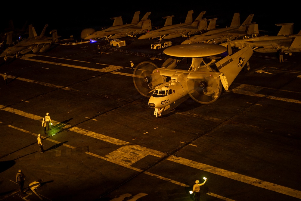 A U.S. Navy E-2 Hawkeye airborne early warning aircraft (bureau number 604) is taxiing on an aircraft carrier flight deck at night, with both turboprop engines running and propellers spinning. Flight deck crew members are visible using illuminated wands to guide the aircraft. Multiple fighter jets, likely F/A-18 Hornets or Super Hornets, are parked in the background along with ground support vehicles.