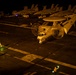 A U.S. Navy E-2 Hawkeye airborne early warning aircraft (bureau number 604) is taxiing on an aircraft carrier flight deck at night, with both turboprop engines running and propellers spinning. Flight deck crew members are visible using illuminated wands to guide the aircraft. Multiple fighter jets, likely F/A-18 Hornets or Super Hornets, are parked in the background along with ground support vehicles.