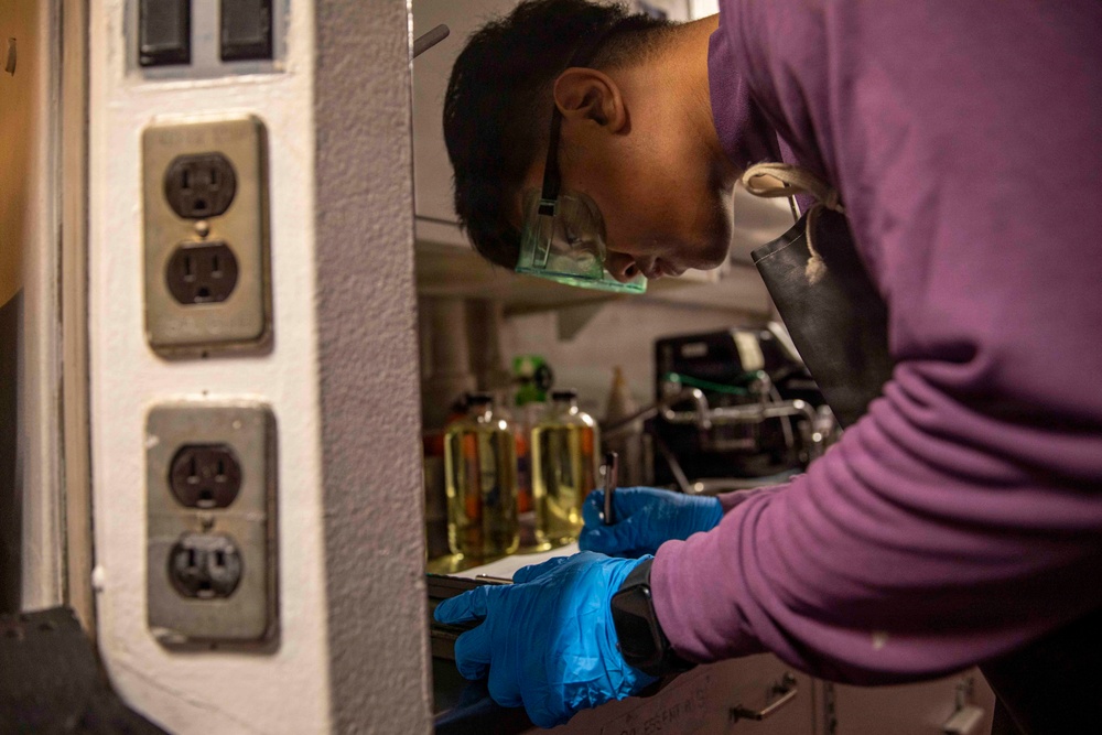 A person wearing blue nitrile gloves, safety goggles, and a purple long-sleeve shirt is working at what appears to be a laboratory or technical workspace. They are bent over a surface, handling tools or equipment with focused attention. The background contains yellow liquid-filled containers and various lab or technical equipment.