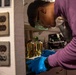 A person wearing blue nitrile gloves, safety goggles, and a purple long-sleeve shirt is working at what appears to be a laboratory or technical workspace. They are bent over a surface, handling tools or equipment with focused attention. The background contains yellow liquid-filled containers and various lab or technical equipment.